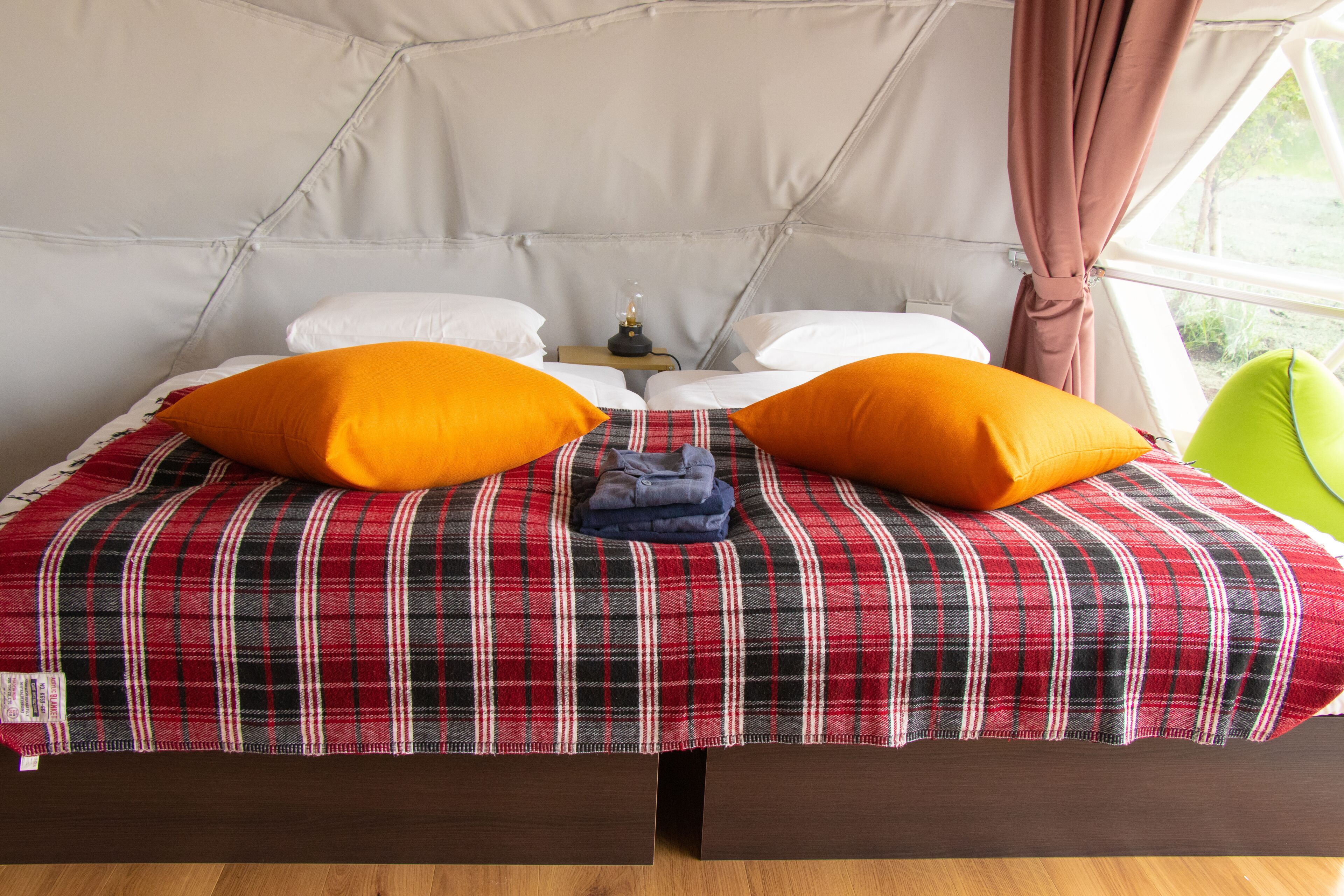 Een bed met een geruit sprei, twee oranje kussens en een groene stoel in een tentachtige ruimte.