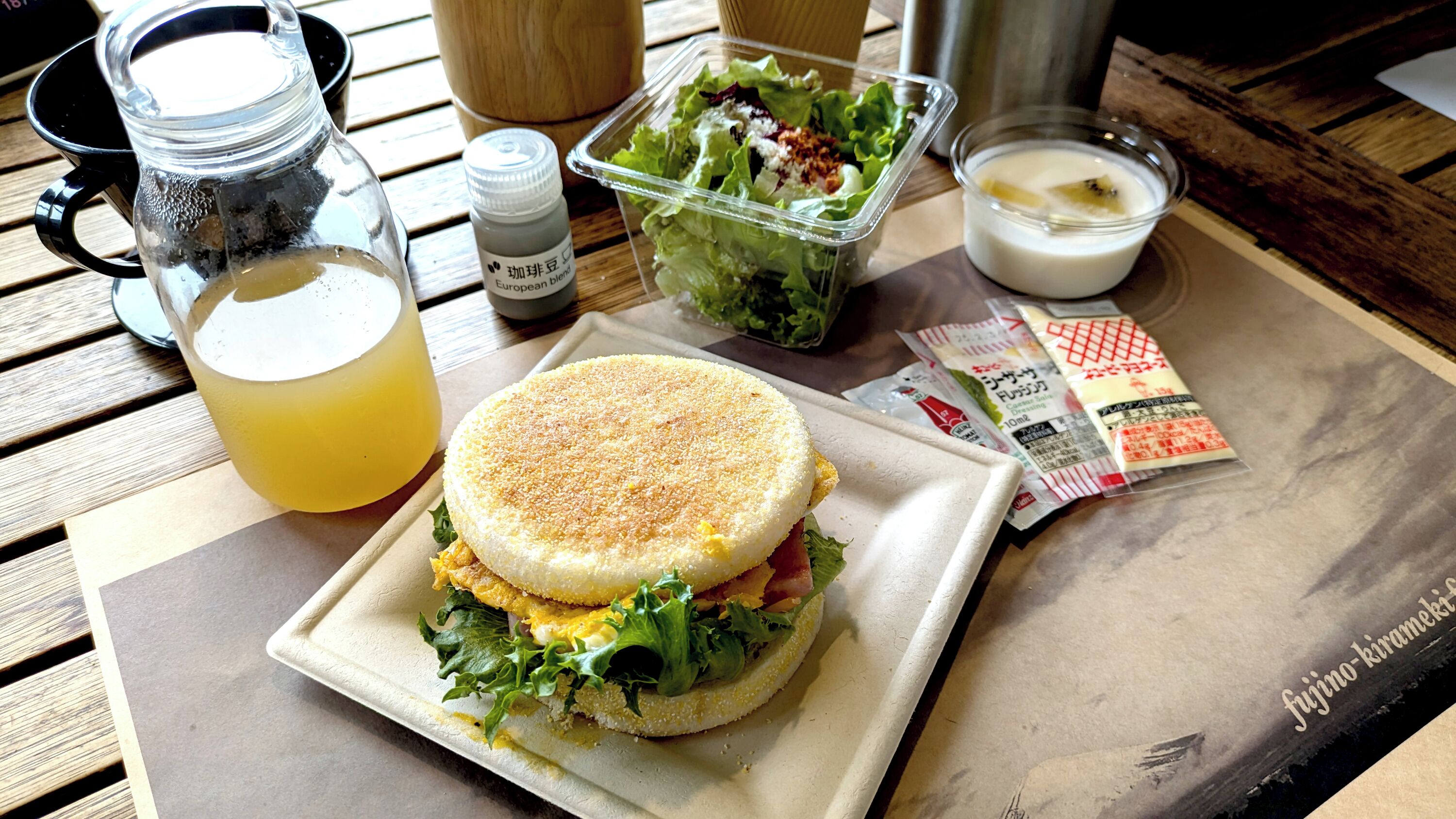A sandwich on a plate, a pitcher of lemonade, a salad container, and condiments on a wooden table.