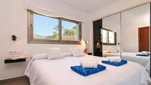 Habitación de hotel con una cama grande, una ventana que ofrece vistas a palmeras, una mesita de noche con lámpara y un espejo que refleja la habitación.