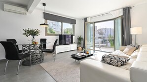 Una sala de estar moderna con una mesa de comedor de vidrio, sillas negras, un sofá blanco y un ventanal con vistas a la ciudad.