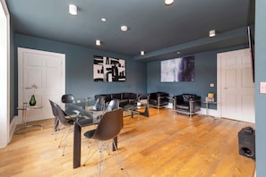 A modern living room with a glass dining table, black leather sofas, and a large screen wall.