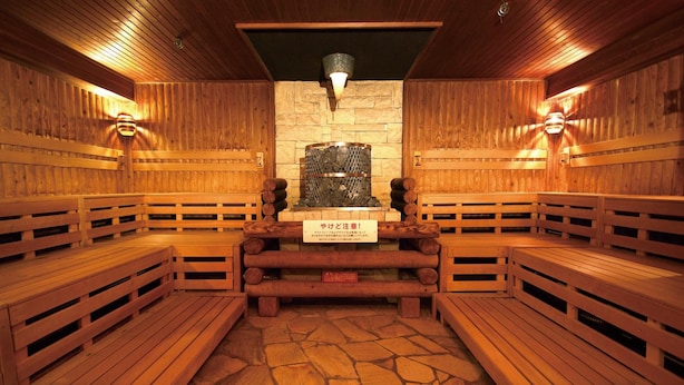 A sauna with wooden benches arranged in a semi-circle, a stone floor, and a central stone wall with a metal container. - Nikoh Capsule Hotel Refre - Hostel - Caters to Men (Sapporo)
