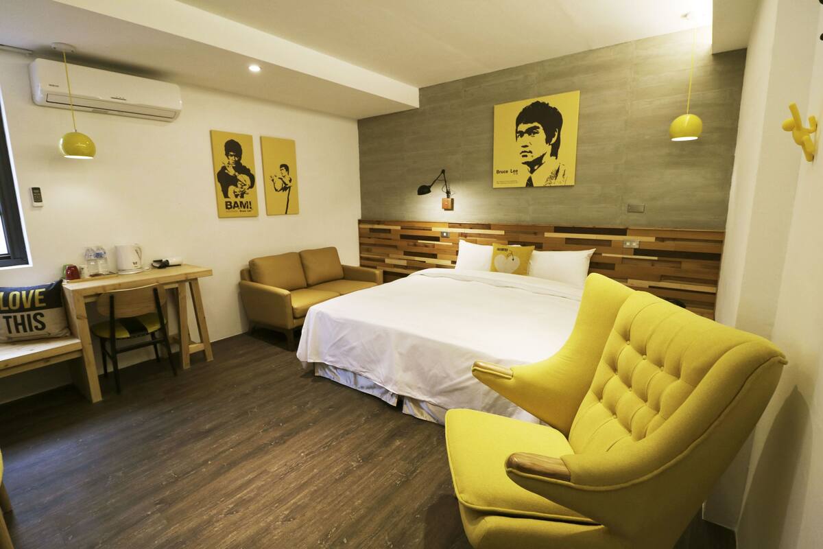Habitación de hotel con una cama grande, un escritorio con silla, un sillón amarillo y dos carteles en la pared.