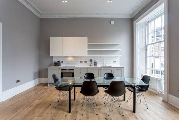 A modern kitchen with a glass dining table and chairs, white cabinetry, and a large window.