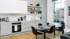 Ett modernt kök med vita skåp, en matplats med ett glasbord och svarta stolar, och en utsikt över byggnader genom ett stort fönster.