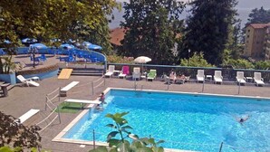 A swimming pool with lounge chairs and umbrellas, surrounded by greenery and buildings. - Selva 1 lake view apartment in Ghiffa with pool (Ghiffa)