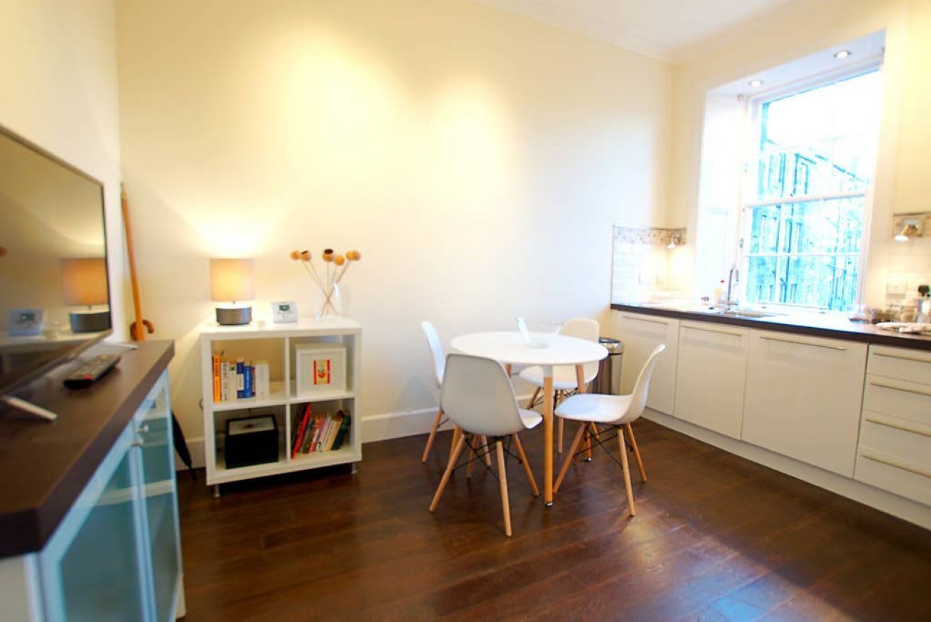 A modern kitchen with white cabinets, a round dining table with white chairs, a built-in oven, and a window with a view of a building.