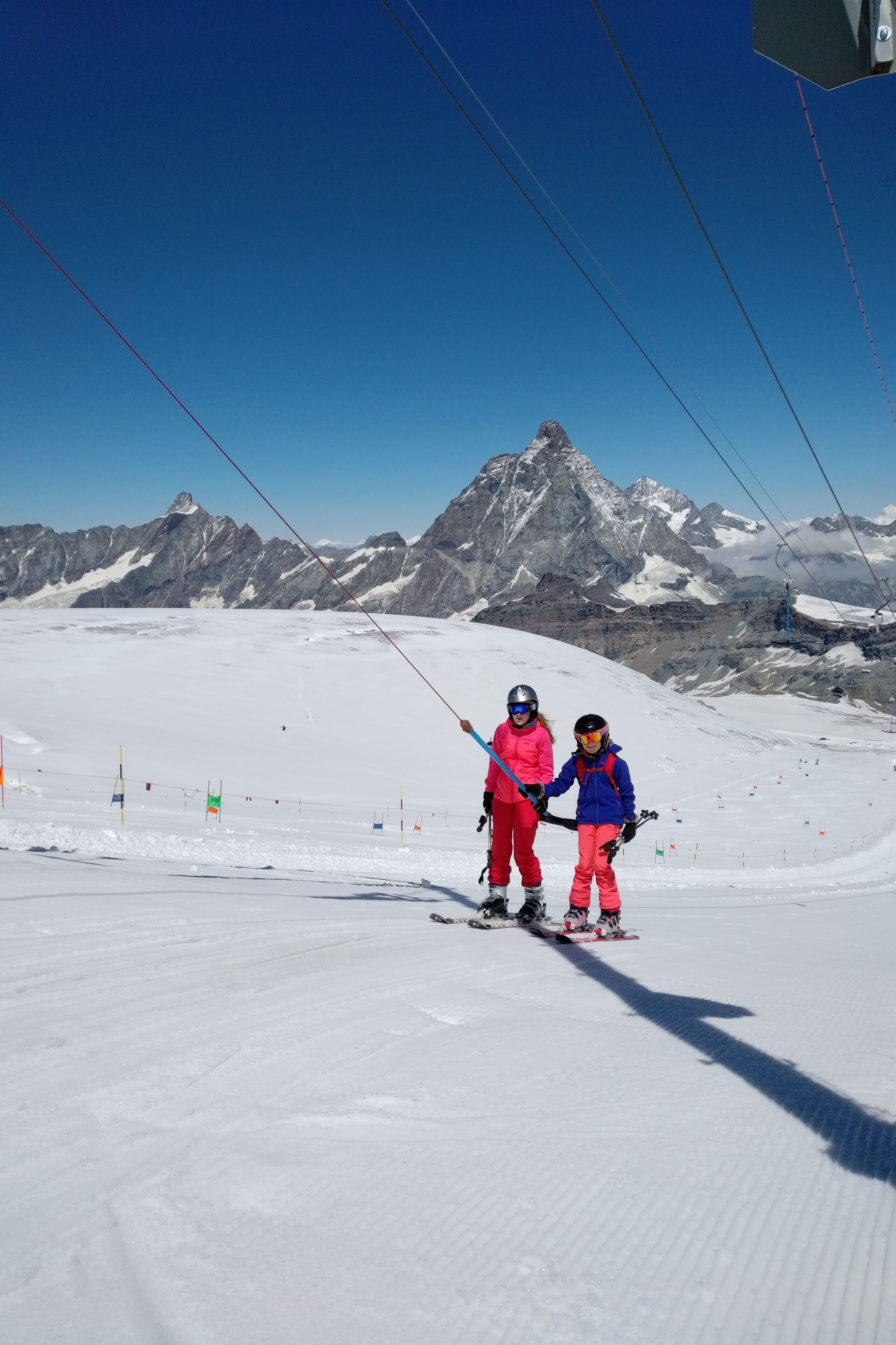 Pista nevada para esquiar con dos esquiadores en primer plano y una cadena montañosa al fondo.
