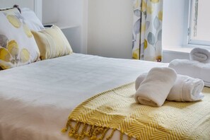 A neatly made bed with a white bedspread, a yellow macrame throw, and two rolled towels on the side.