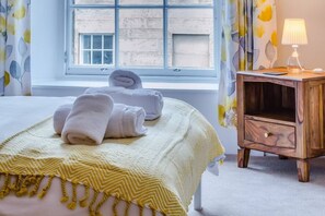 A neatly made bed with rolled towels, a wooden nightstand with a lamp, and a window with a view of a building.