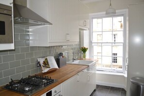 Une cuisine avec des meubles blancs, une cuisinière à gaz, une fenêtre donnant sur des immeubles et un évier.
