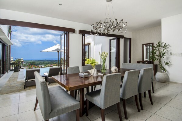 A dining area with a wooden table and grey upholstered chairs, large windows offering a view of the outdoors, and a modern chandelier.