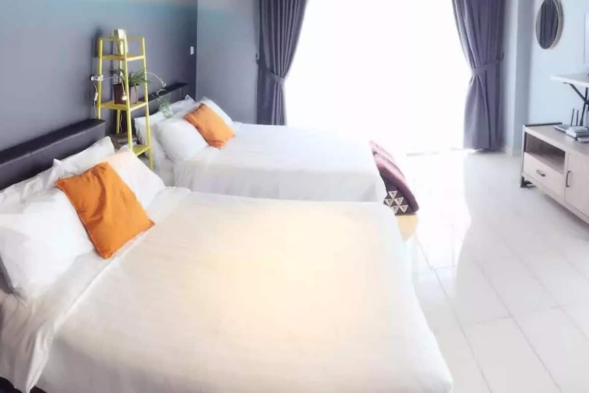 Zwei Betten mit weißen Laken und orangefarbenen Kissen, ein kleiner Tisch mit einer Lampe und ein Regal mit Pflanzen.