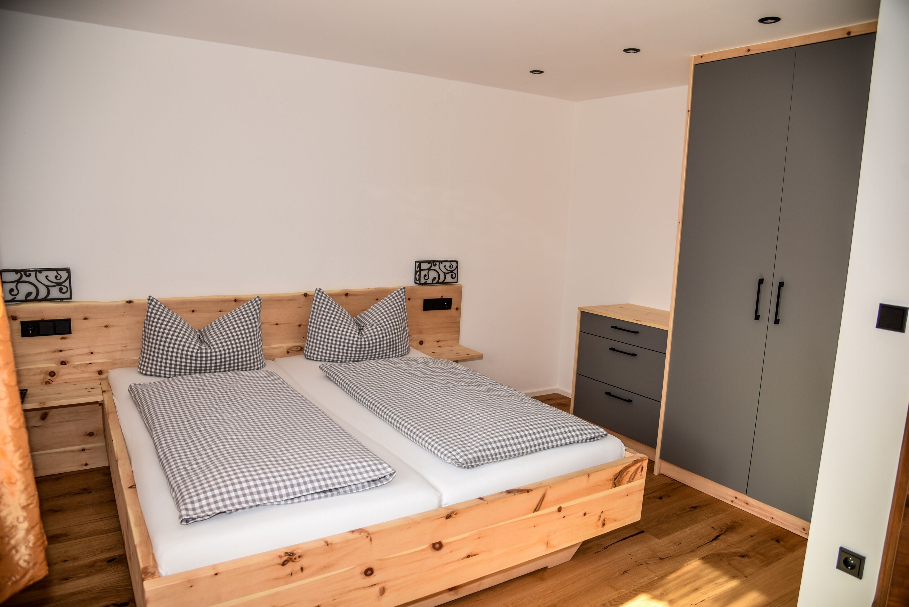 Una cama individual con ropa de cama a cuadros, cabecera de madera, mesitas de noche con adornos y un armario gris.