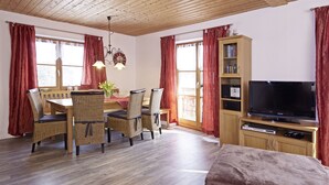 Una sala de estar acogedora con techo de madera, un comedor con sillas de mimbre, un televisor sobre un mueble de madera y una estantería.