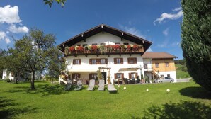 A two-story building with a balcony adorned with flowers, a lawn with outdoor seating, and a car parked nearby. - Ferienwohnung im Erdgeschoss 45 qm, Schlafzimmer, Wohn-/Schlafraum, separate Küche, 4 Sterne, max. 4 Personen (Anger)