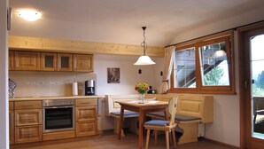 A kitchen with wooden cabinets, a dining table with chairs, a coffee machine, and a window with a view of greenery.