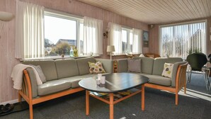 Ein gemütliches Wohnzimmer mit einer Couch, einem Couchtisch und einem Sessel. Es gibt Fenster mit Vorhängen, eine Lampe und ein gerahmtes Bild an der Wand.