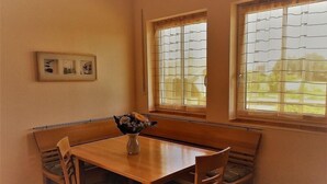 Uma área de refeições com mesa de madeira e bancos, duas janelas com persianas e um quadro na parede.