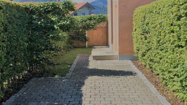 Um caminho pavimentado que leva a um edifício com cerca de madeira e um conjunto de sebes bem cuidadas.
