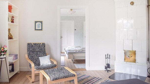 A bright, well-lit room with a wooden floor, a patterned armchair, a small table, and a bookshelf. There is a door leading to another room with a bed and a white wall with a decorative element. - Nice home in Växjö with kitchen (Växjö)