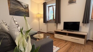 A living room with a sofa, a glass coffee table, a TV on a wooden stand, a lamp, and a window with curtains. - Apartment no. 4 Amara for 1-3 pers., 42 m², 1 sep. bedroom. (Bischofswiesen)