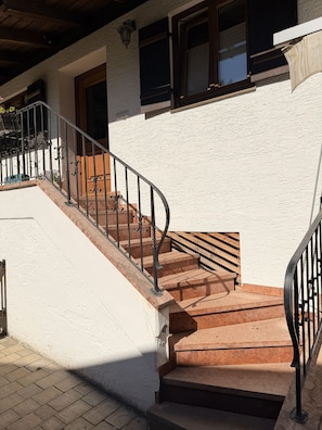 A staircase with a wooden handrail and metal balusters leading to a building with white walls and wooden doors.
