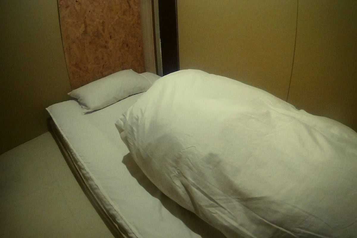 Une petite chambre avec un lit simple, dotée d’une tête de lit en bois et d’un matelas blanc uni.