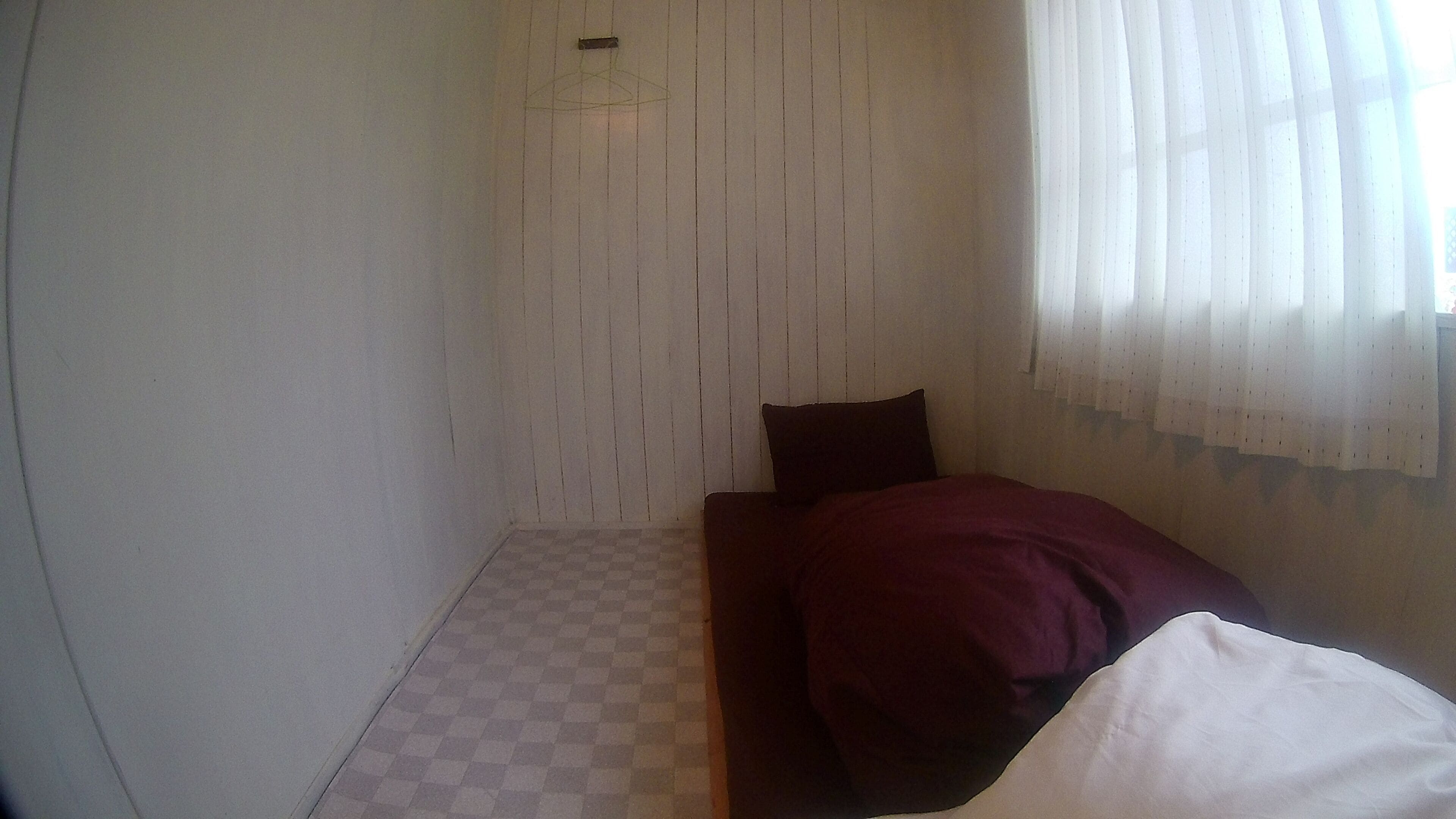 Una habitación pequeña, con poca luz, con piso de tablones, una cama individual con una manta oscura y una ventana con cortinas translúcidas.