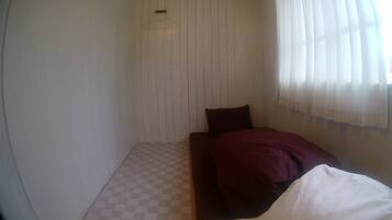 Una habitación pequeña, con poca luz, con piso de tablones, una cama individual con una manta oscura y una ventana con cortinas translúcidas.