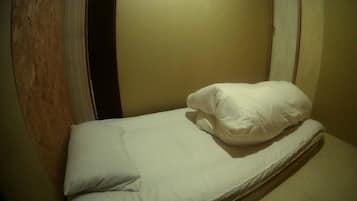 A small, single-bed room with a wooden door frame, a plain wall, and a white mattress with a single pillow.