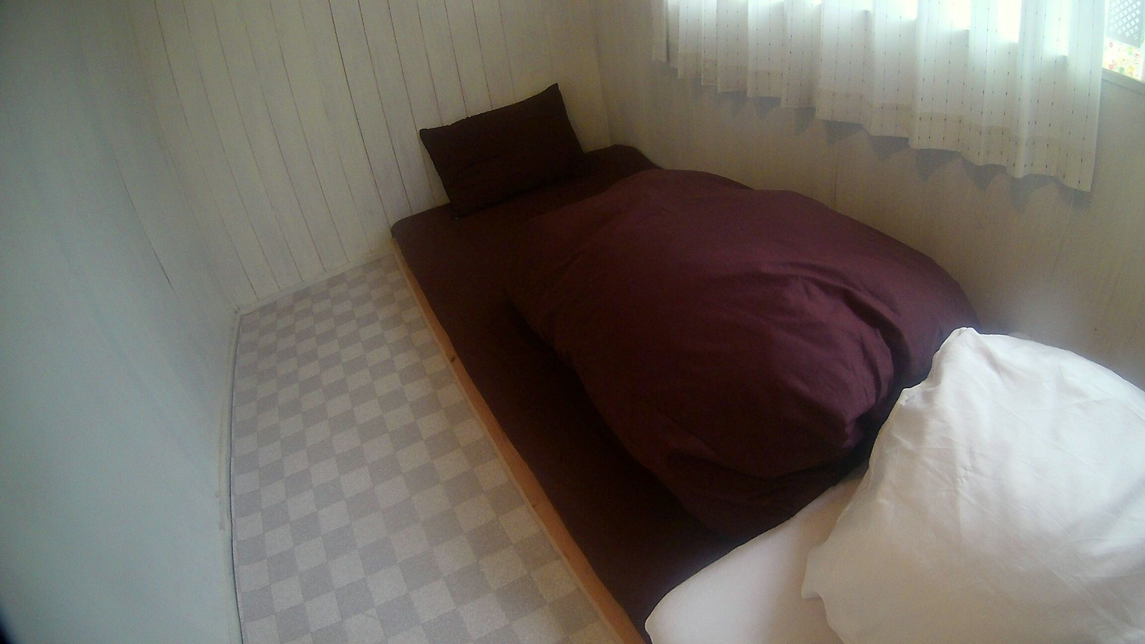 A small, single-bed room with a checkered floor, a dark brown headboard, and white curtains.
