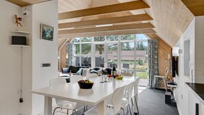 Eine moderne Küche mit weißen Schränken, eine Essecke mit weißem Tisch und Stühlen und große Fenster mit Blick ins Freie.
