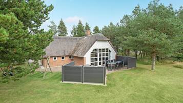 Une petite maison au toit de chaume, dotée d’une grande fenêtre et d’un patio aménagé avec des meubles d’extérieur.