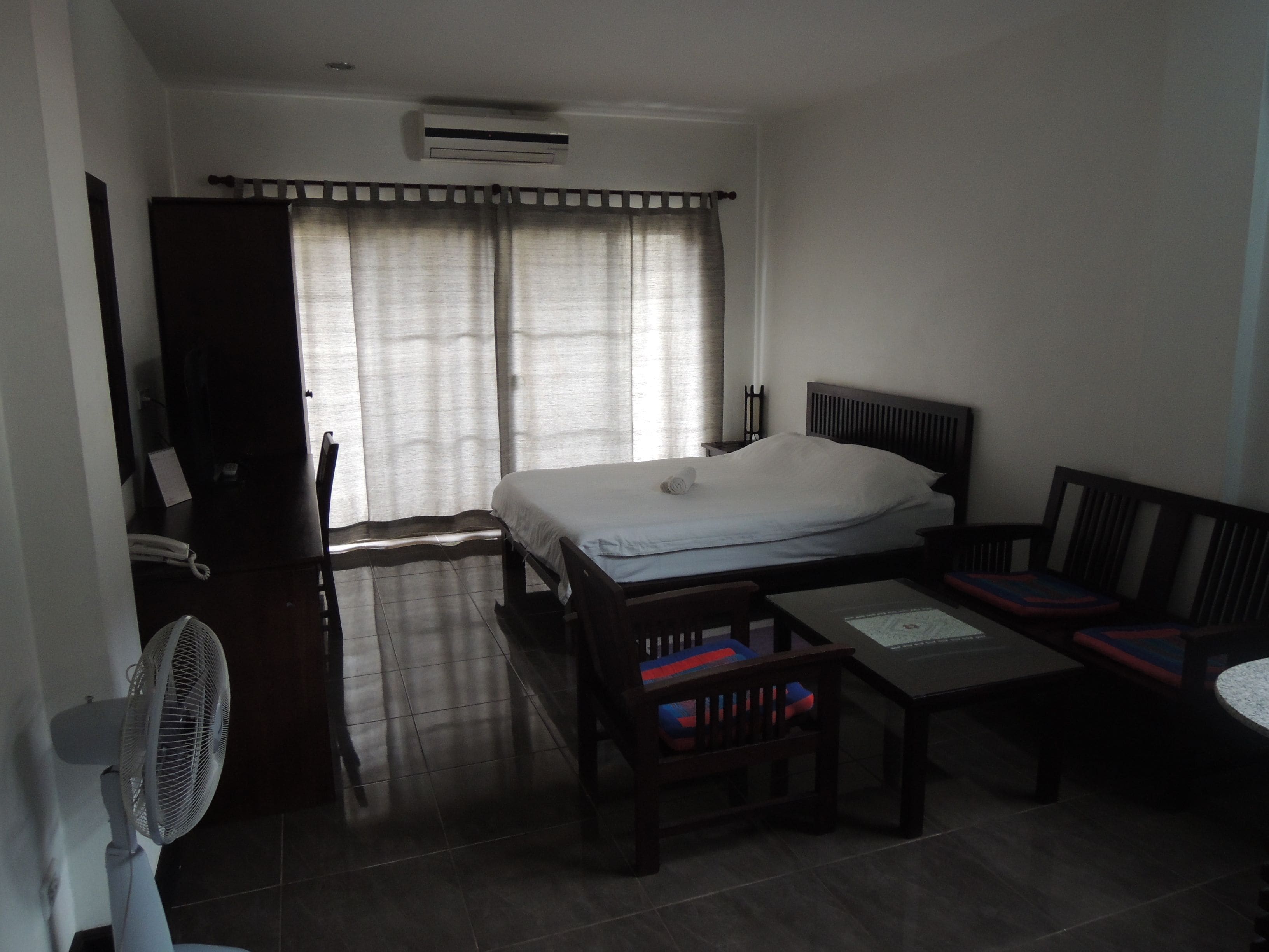 Een hotelkamer met een bed, een bureau, een stoel, een tafel, een ventilator en een raam met gordijnen.