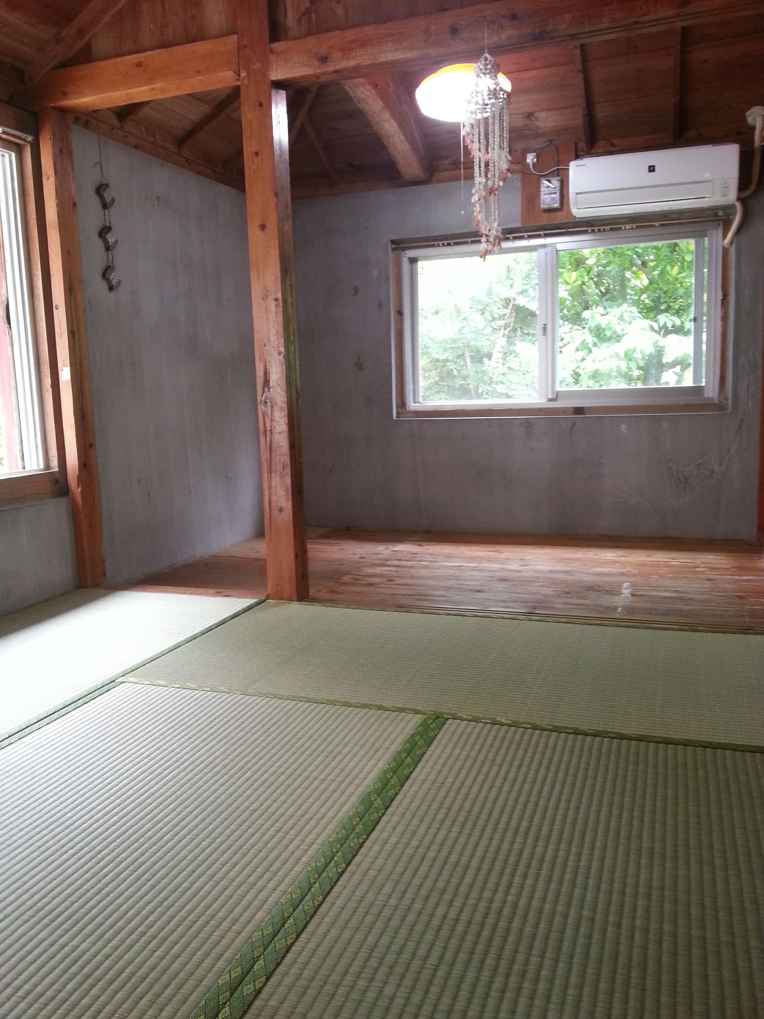 A room with tatami mats, a wooden ceiling, a window with a view of trees, and an air conditioning unit.