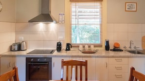 A kitchen with a wooden table, chairs, a stove, a sink, and a window with blinds. - Rubys Cottage (Penola)