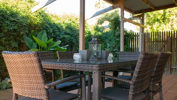 A covered patio with wicker furniture and a glass table, surrounded by lush greenery. - Rubys Cottage (Penola)
