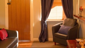 A living room with a dark-colored sofa, a matching armchair, a wooden door, and a window with dark curtains. - Rubys Cottage (Penola)