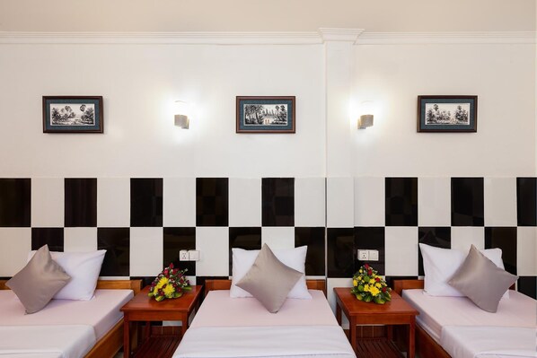 A hotel room with two beds, each with a pillow and a small table with a vase of flowers. There are three framed pictures on the wall above the beds. - Botoum Hostel (Siem Reap)