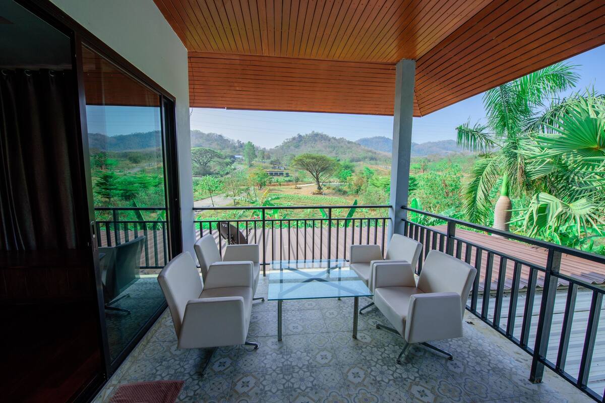 A balcony with a glass table and four chairs, overlooking a lush landscape with trees and hills.