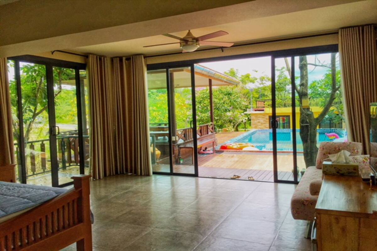 A room with a wooden bed, a desk with a chair, and a view of a swimming pool and greenery outside.