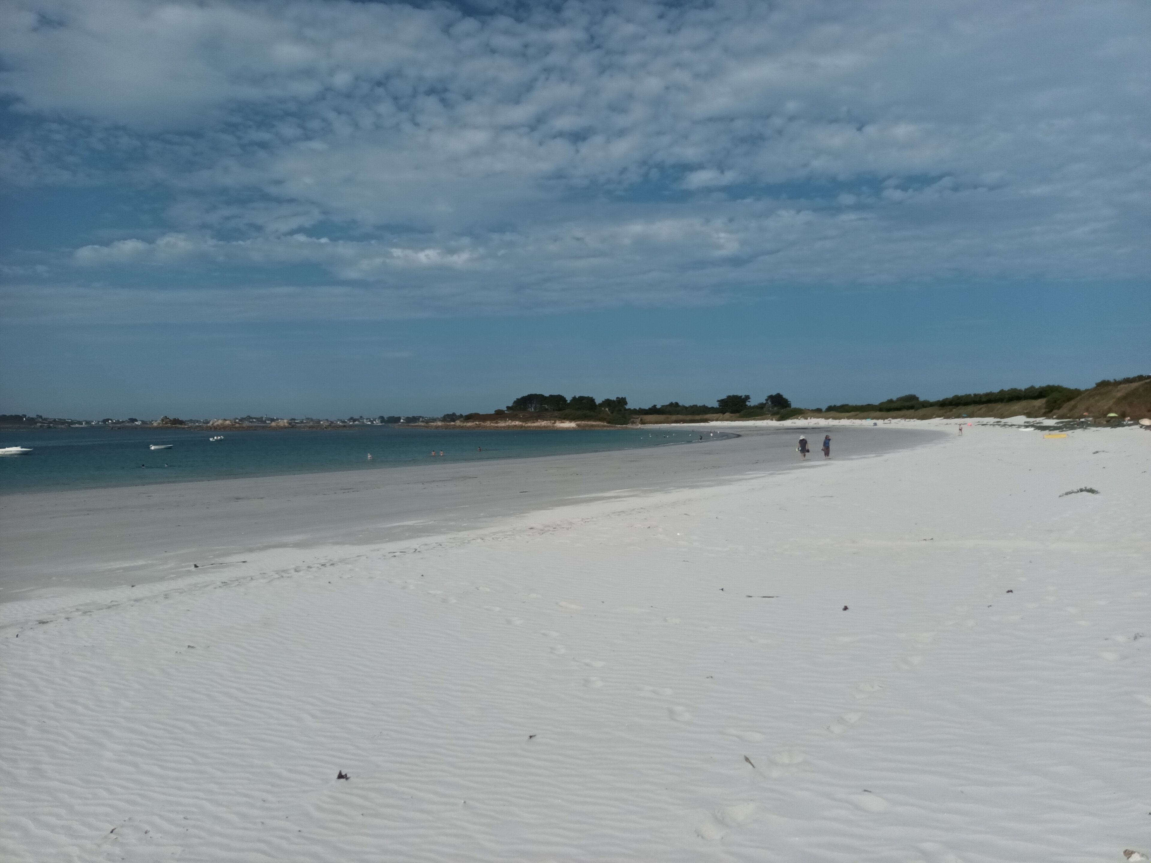 Una playa con una amplia orilla arenosa, agua tranquila y algunas personas caminando.
