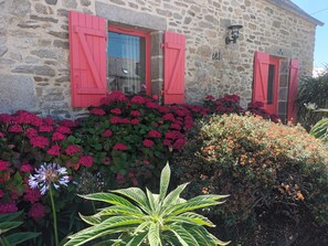 Un edificio de piedra con contraventanas rojas y un jardín con diversas plantas y flores.