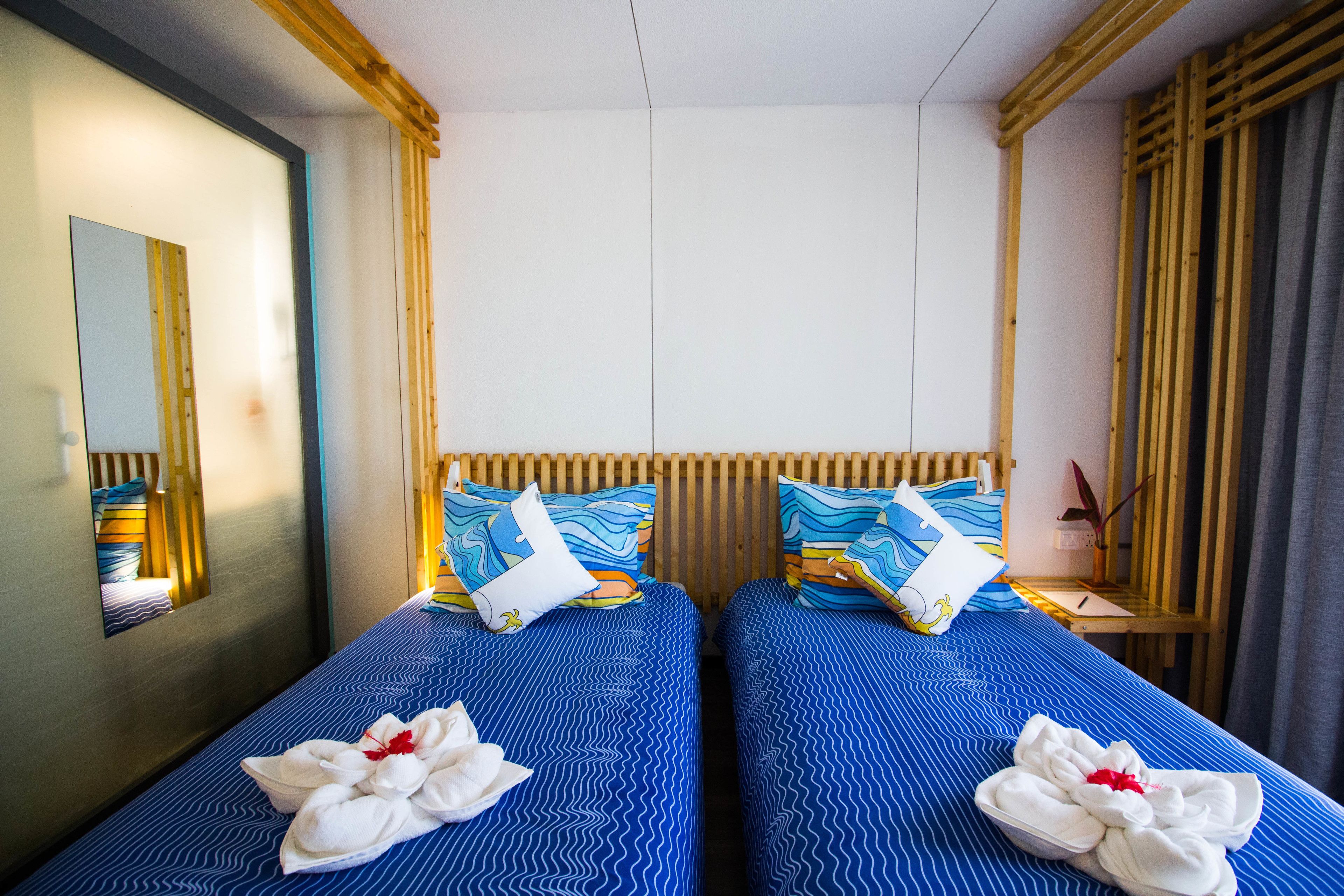 Hotelkamer met twee bedden, elk met een blauw beddekleed en een witte handdoek met een rode bloem. Een houten hoofdeinde, een spiegel en een klein tafeltje met een plantje.