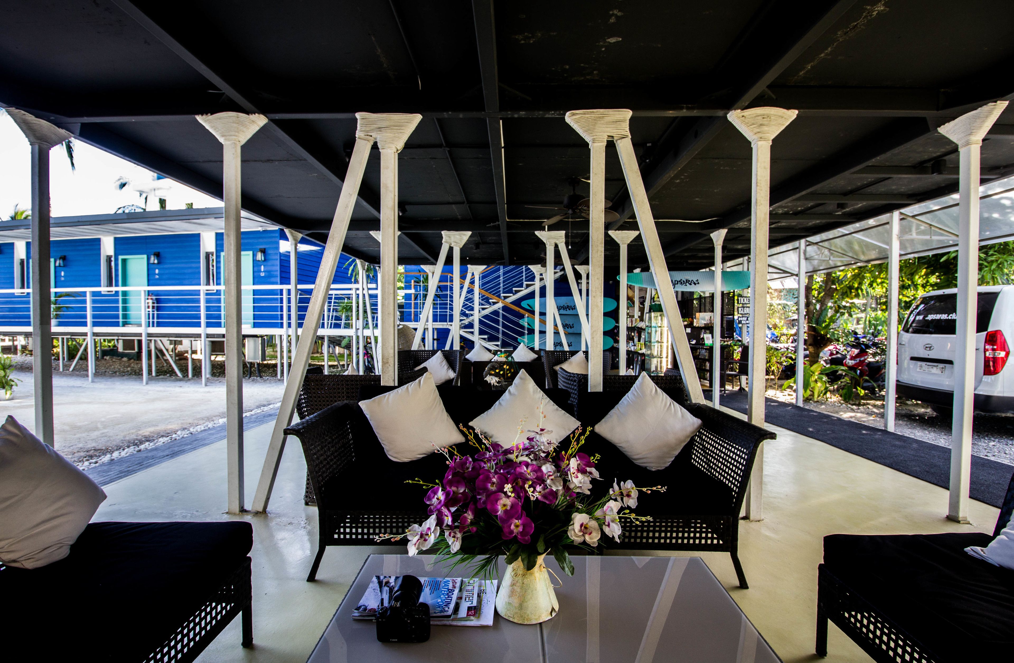 A covered outdoor seating area with a sofa and chairs, a table with a vase of flowers, and a view of a blue building in the background.
