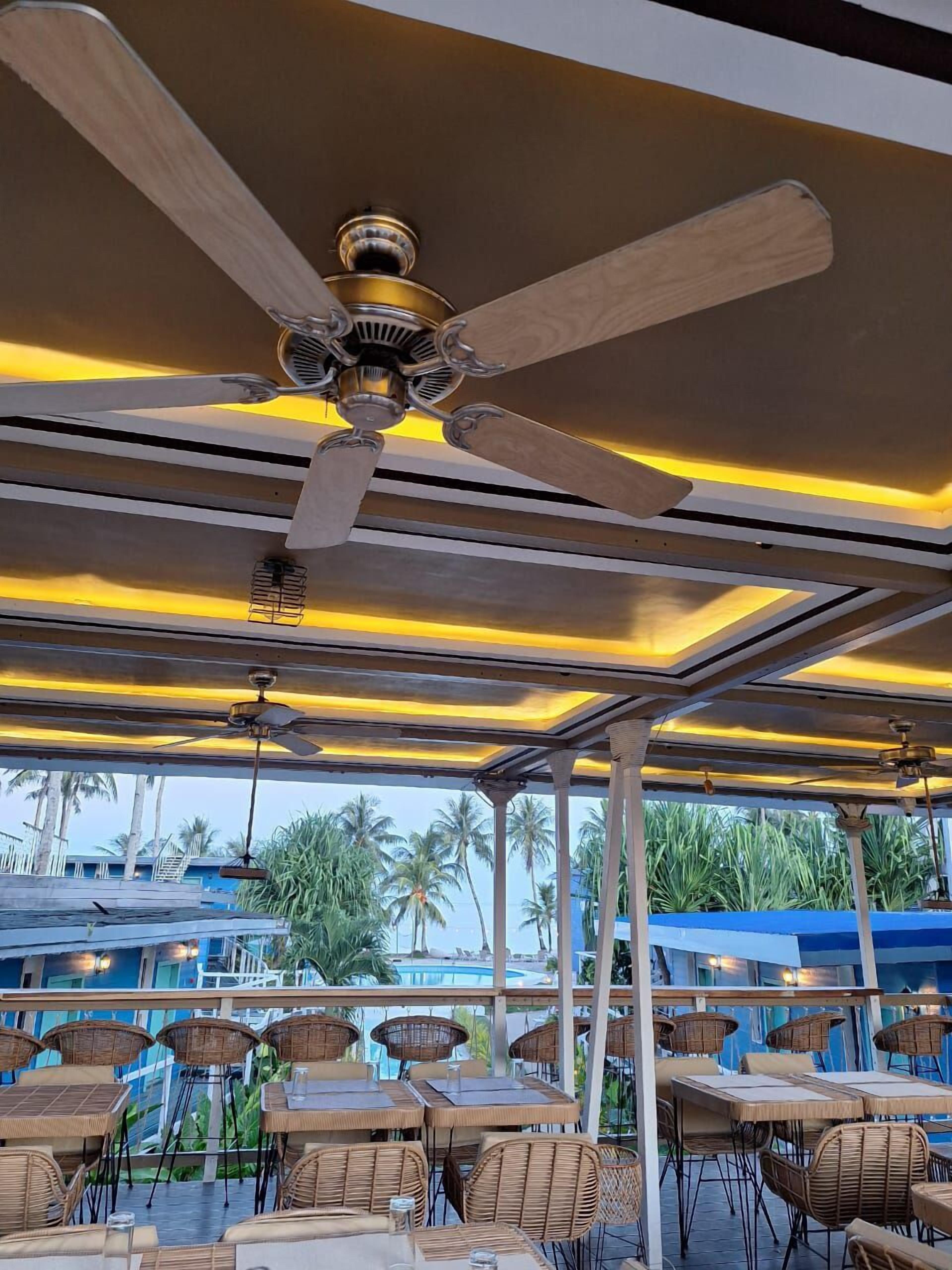 An outdoor seating area with tables and chairs, a ceiling fan, and a view of palm trees and a pool.