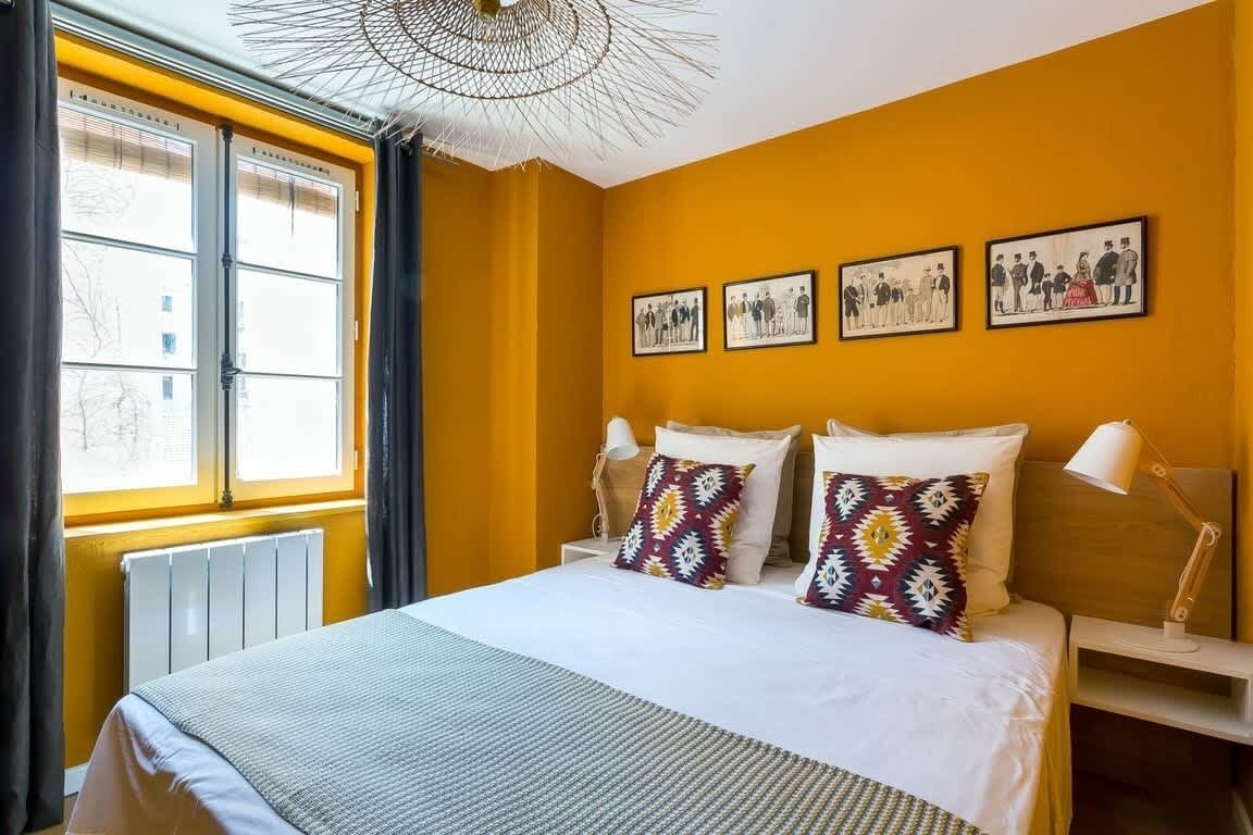 A hotel room with a large bed, a window with blinds, a radiator, and three framed pictures on the wall.