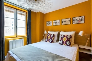 Une chambre d’hôtel avec un grand lit, une fenêtre avec des stores, un radiateur et trois tableaux encadrés accrochés au mur.