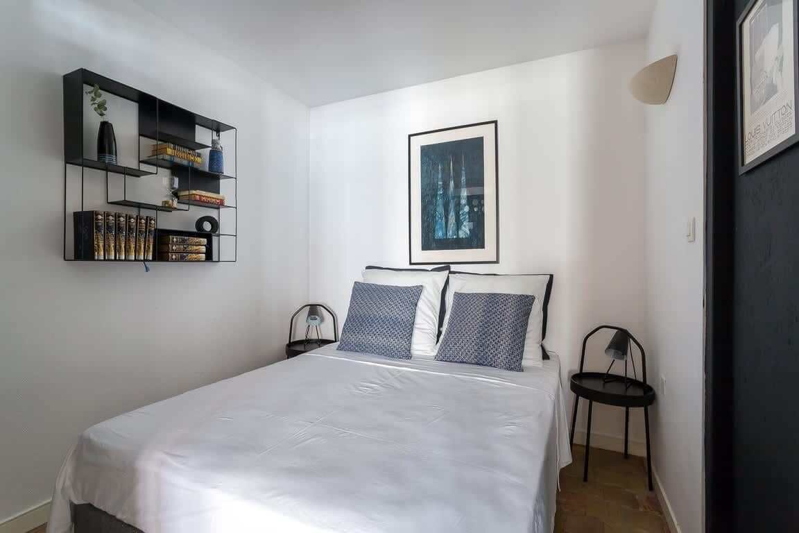 A bedroom with a bed, a wall-mounted shelf with books, a framed picture, and two chairs.
